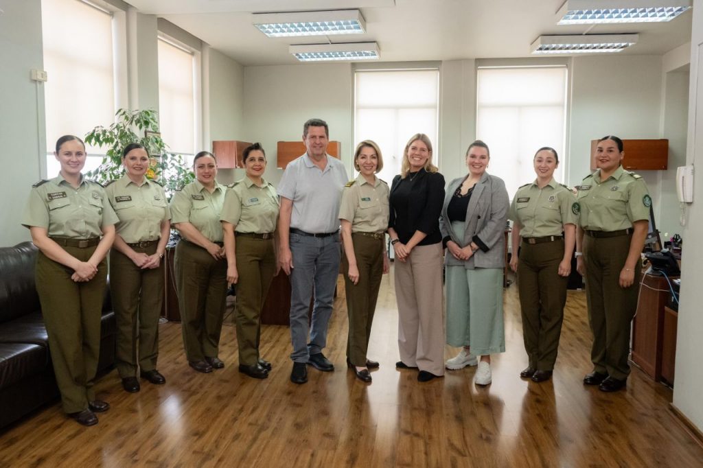 The picture shows a group of people, mostly women and one man, posing for a camera in a bright room. Except of two women and one woman, the women are police officers and wear light green and dark green police uniforms.