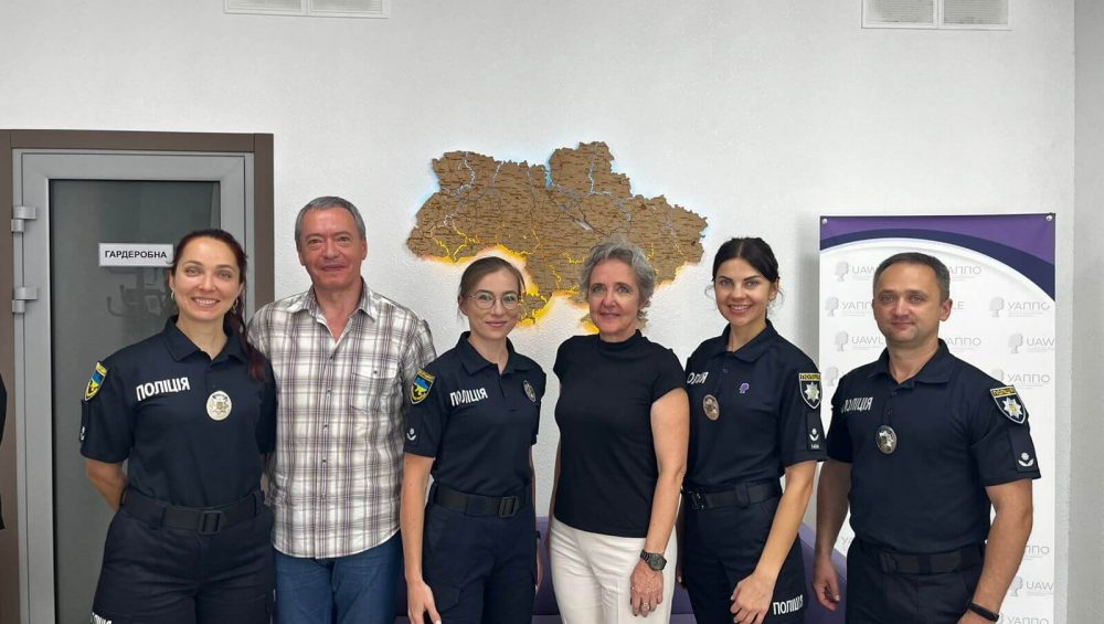 The photo shows three female officers and one male officer of the Ukrainian National Police together with two international guests. They are standing in a row in front of a wall with an illuminated map of Ukraine. Everyone is smiling at the camera, creating a professional yet friendly atmosphere.