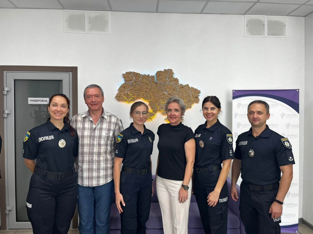 The photo shows three female officers and one male officer of the Ukrainian National Police together with two international guests. They are standing in a row in front of a wall with an illuminated map of Ukraine. Everyone is smiling at the camera, creating a professional yet friendly atmosphere.