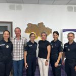 The photo shows three female officers and one male officer of the Ukrainian National Police together with two international guests. They are standing in a row in front of a wall with an illuminated map of Ukraine. Everyone is smiling at the camera, creating a professional yet friendly atmosphere.