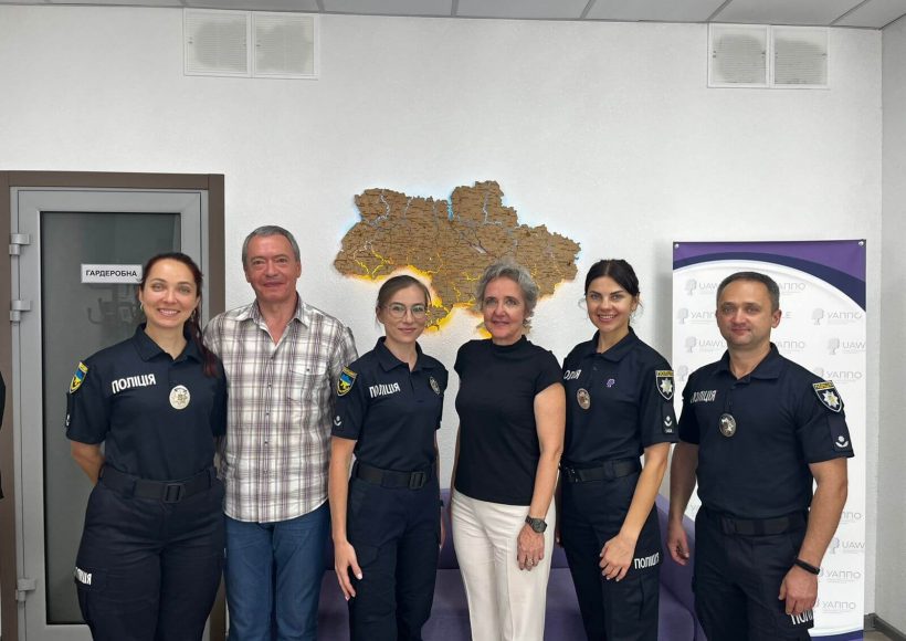 The photo shows three female officers and one male officer of the Ukrainian National Police together with two international guests. They are standing in a row in front of a wall with an illuminated map of Ukraine. Everyone is smiling at the camera, creating a professional yet friendly atmosphere.