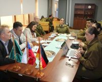 Briefing at Carabineros office; desk flags and laptop in foreground as officers and GS-Foundation delegates confer.